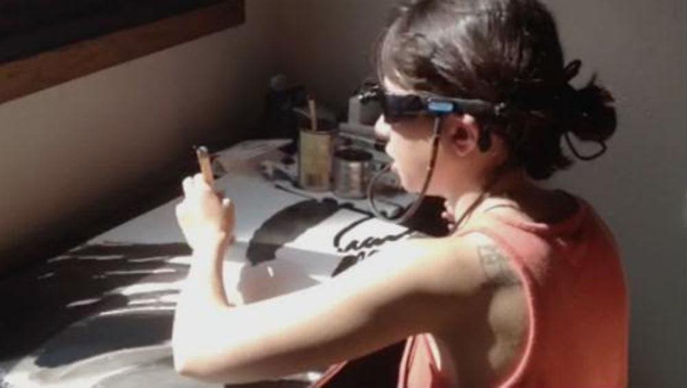 Emilie Gossiaux sits at a table and paints with black paint while wearing the Brainport Visions Device.