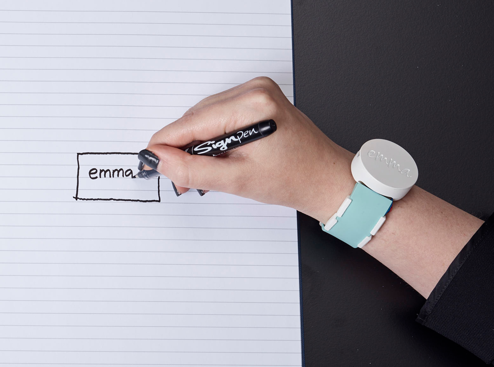 A woman wearing watch-like device, The Emma, on her wrist writes the name Emma on a sheet of lined paper.