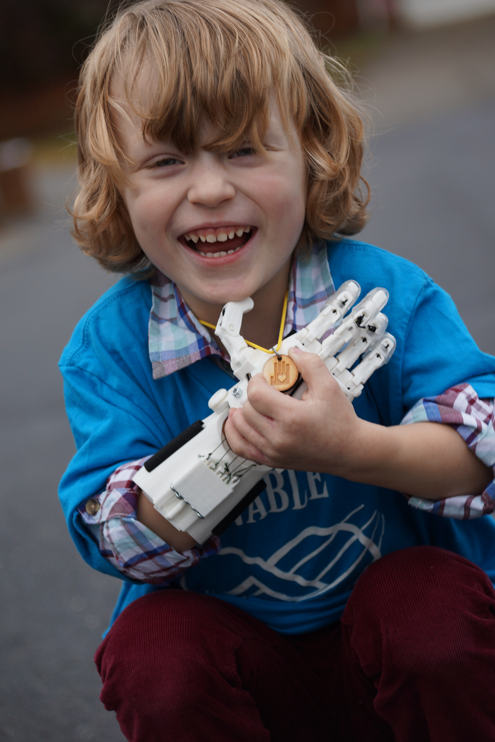 A laughing young boy squats, holding his right, 3D printed hand to his chest and holding an e-Nable necklace with his left hand.